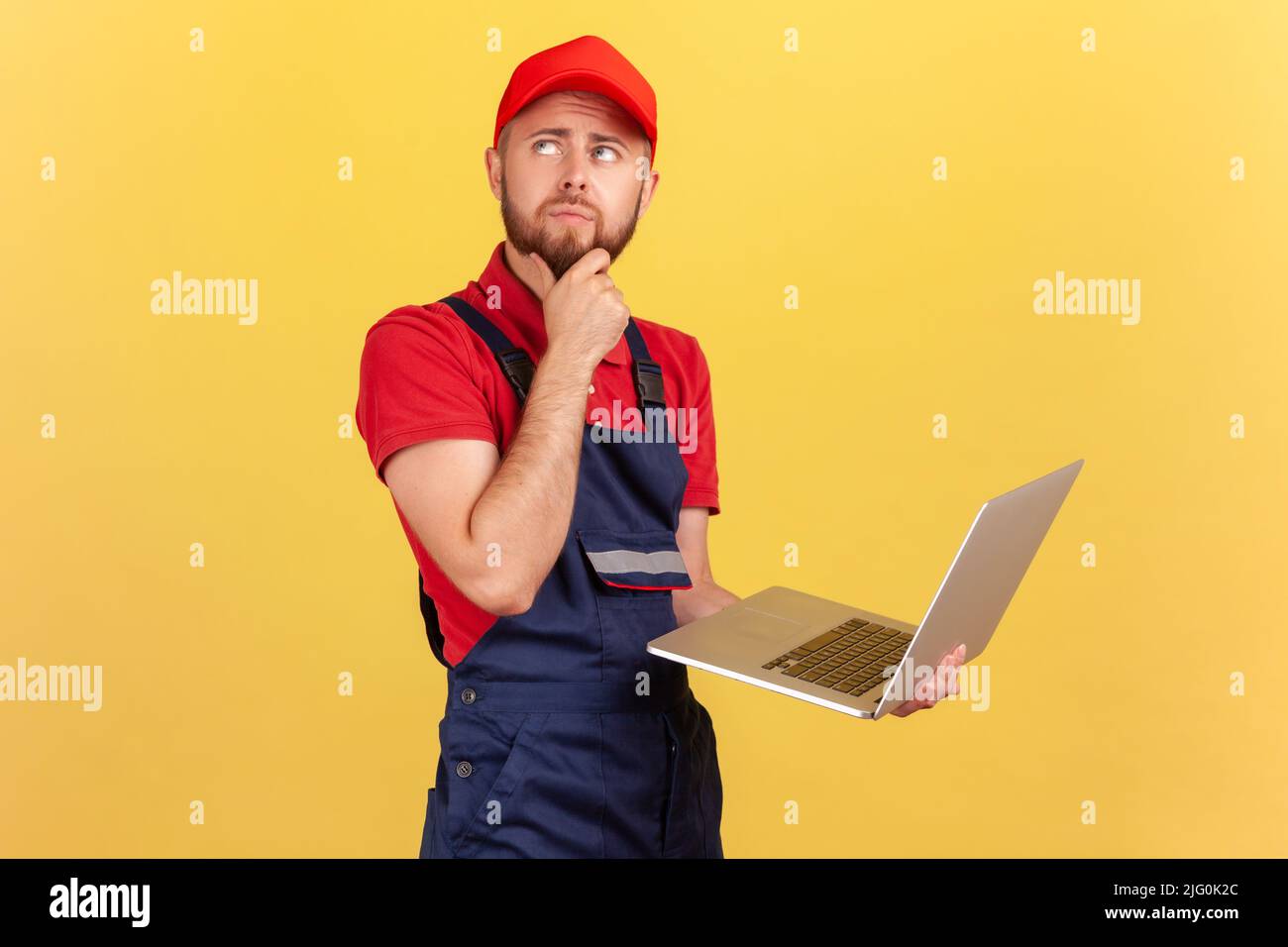 Porträt eines bärtigen, nachdenklichen Arbeiters, der auf dem Laptop steht und arbeitet, Bestellungen entgegennimmt, online unterstützt, das Kinn hält, über eine Lösung nachdenkt. Innenaufnahme des Studios isoliert auf gelbem Hintergrund. Stockfoto