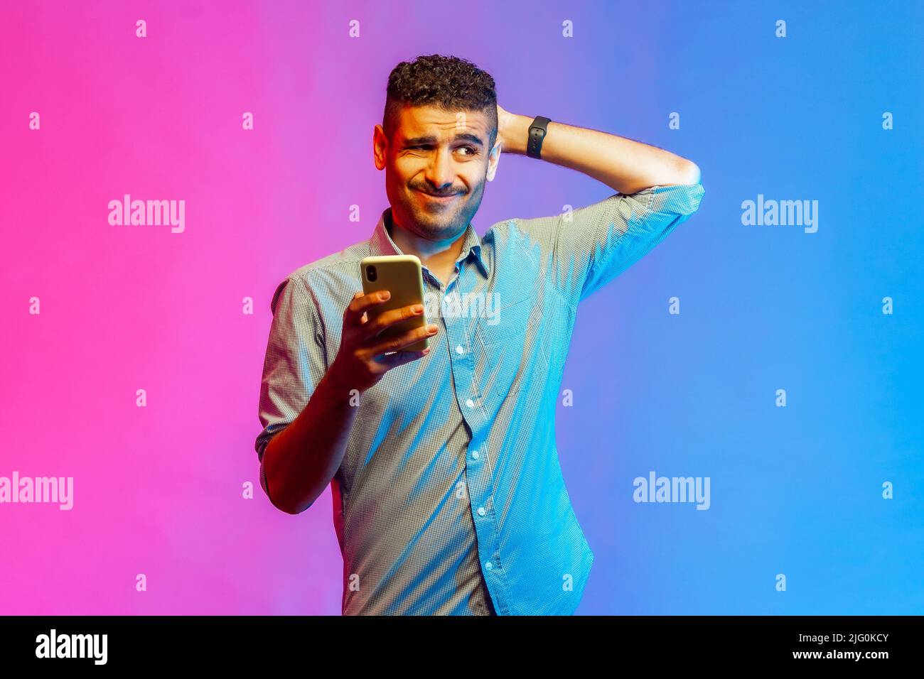 Portrait eines Mannes im Hemd kratzender Kopf, der das Smartphone in der Hand hält, über Software-Updates nachdenkt, geeignete Tarife wählt. Innenaufnahmen im Studio, isoliert auf farbigem Neonlicht-Hintergrund. Stockfoto