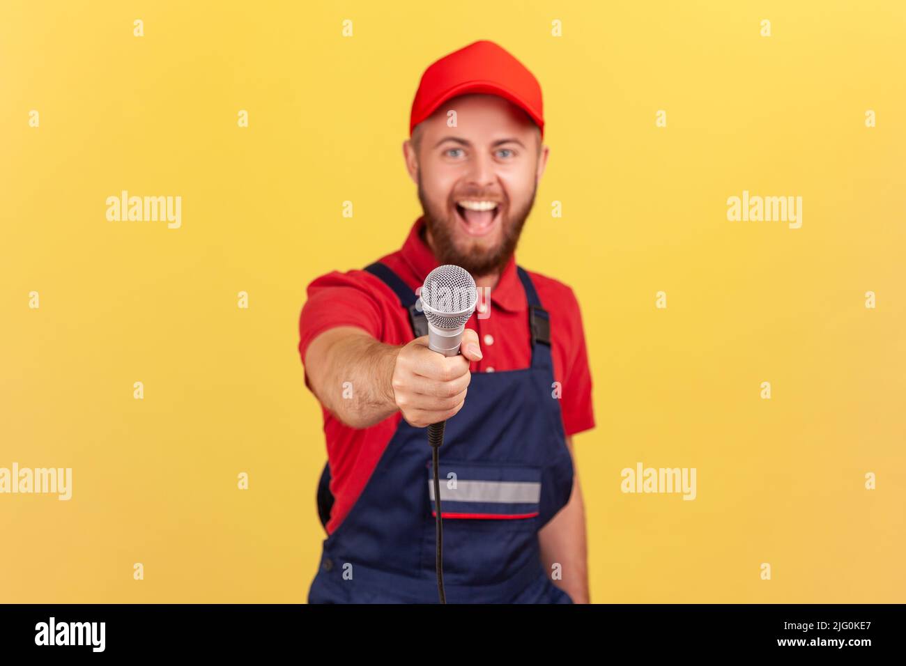 Porträt eines aufgeregten bärtigen Arbeiters in blauer Uniform und Mütze, der stand und ein Mikrofon anbot und die Kamera mit fröhlichem Gesichtsausdruck ansah. Innenaufnahme des Studios isoliert auf gelbem Hintergrund. Stockfoto