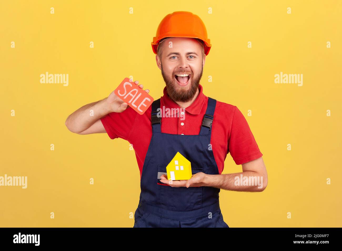 Portrait eines glücklichen lächelnden Bauherrn, der Papierhaus und Karte mit Verkaufsaufschrift hält und anbietet, eine neue Wohnung zu einem niedrigen Preis zu kaufen. Innenaufnahme des Studios isoliert auf gelbem Hintergrund. Stockfoto