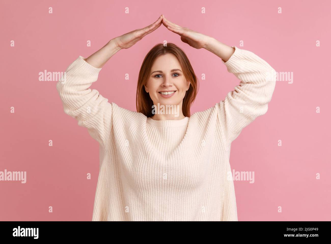 Porträt einer fröhlichen, positiven jungen blonden Frau, die mit den Armen über dem Kopf steht, Dachbewegung, Heimsymbol, weißen Pullover trägt. Innenaufnahme des Studios isoliert auf rosa Hintergrund. Stockfoto