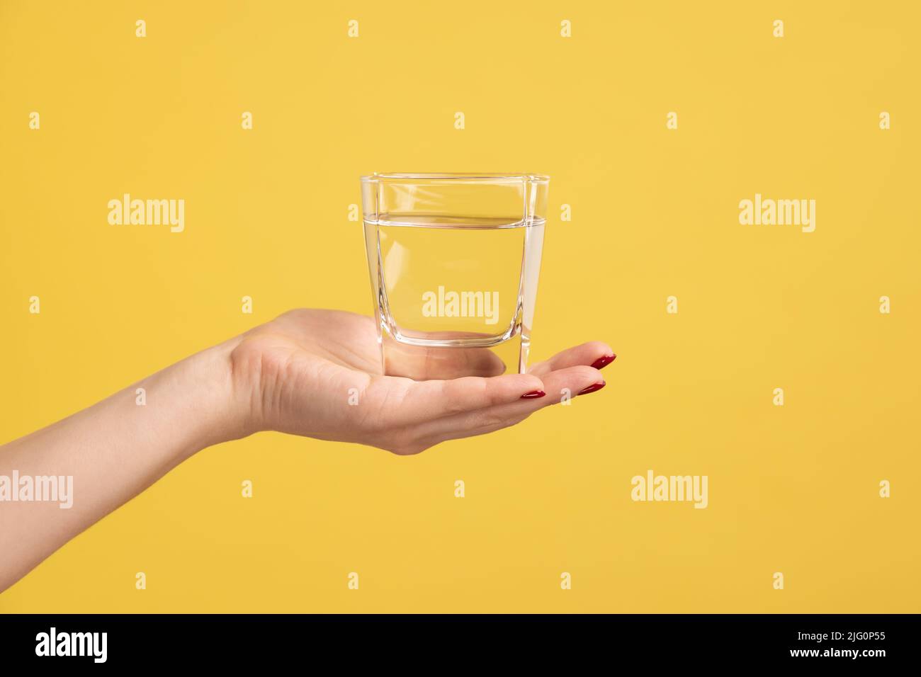 Nahaufnahme Frau Hand hält Glas mit Wasser auf der Handfläche, Werbung für frisches reines Getränk, beste Möglichkeit, um den Wasserhaushalt wiederherzustellen. Innenaufnahme des Studios isoliert auf gelbem Hintergrund. Stockfoto