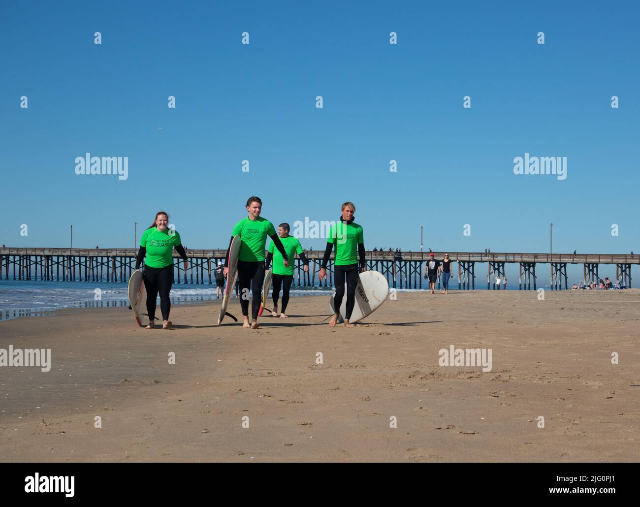 Vier junge Rettungsschwimmer mit grünen Tops und Surfbrettern laufen stolz am Strand von Newport Beach, Kalifornien, USA entlang Stockfoto