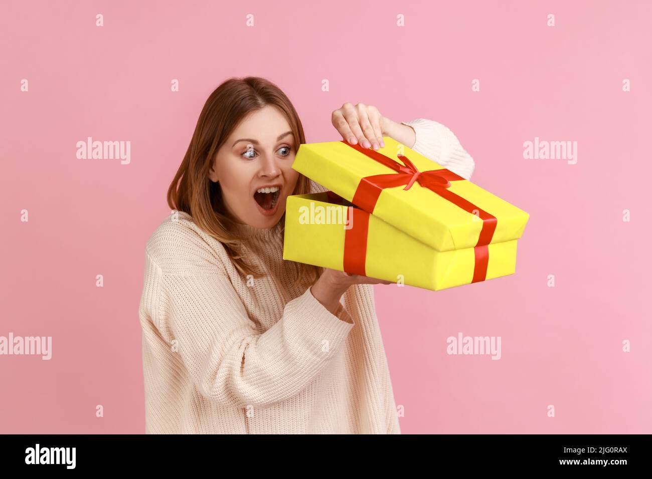 Porträt der aufgeregt jungen erwachsenen blonde Frau in gelb Geschenk-Box suchen, interessiert, was im Inneren, trägt weißen Pullover. Innenaufnahme des Studios isoliert auf rosa Hintergrund. Stockfoto