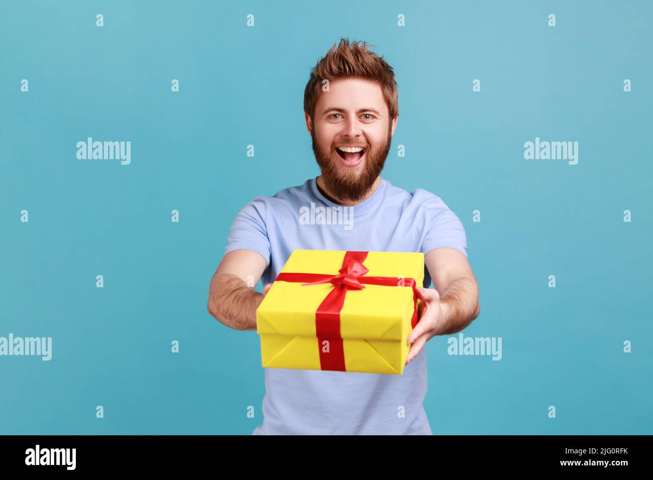 Porträt eines positiven bärtigen Mannes, der gelbe Geschenkbox mit rotem Band an die Kamera gibt und lächelt, das Weihnachtsgeschenk teilt und gratuliert. Innenaufnahme des Studios isoliert auf blauem Hintergrund. Stockfoto