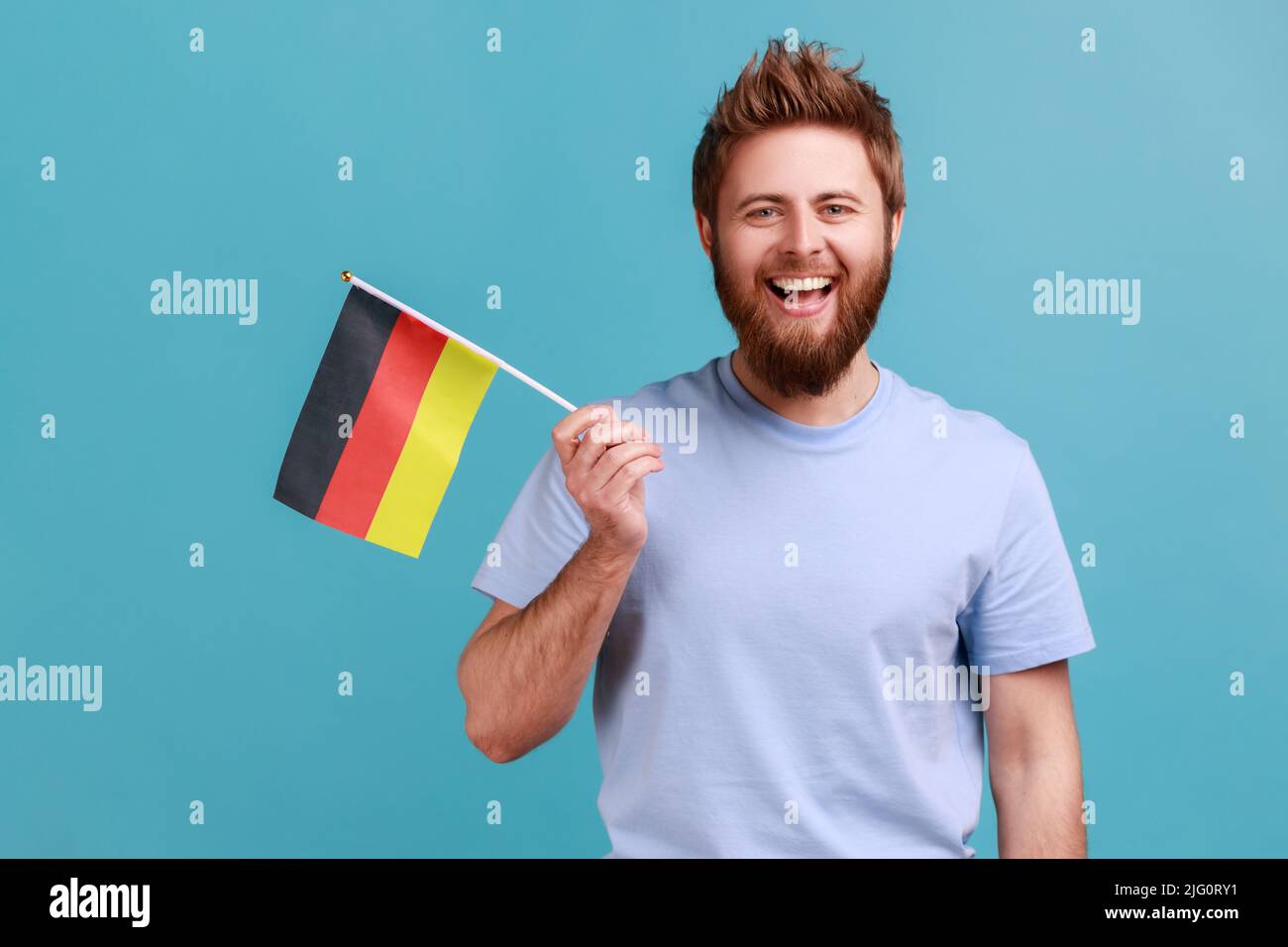 Porträt eines glücklichen, zufriedenen bärtigen Mannes mit der deutschen Flagge, der den Tag Deutschlands - den 3.. Oktober - feiert und positive Emotionen zum Ausdruck bringt. Innenaufnahme des Studios isoliert auf blauem Hintergrund. Stockfoto