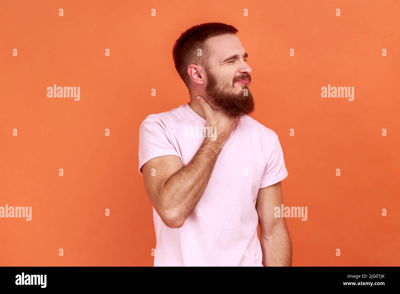 Entzündete Kehle. Porträt eines bärtigen Mannes, der schmerzhaften Hals berührt, Halsschmerzen leidet, Mandelentzündung Symptom, medizinisches Konzept, trägt rosa T-Shirt. Innenaufnahme des Studios isoliert auf orangefarbenem Hintergrund. Stockfoto