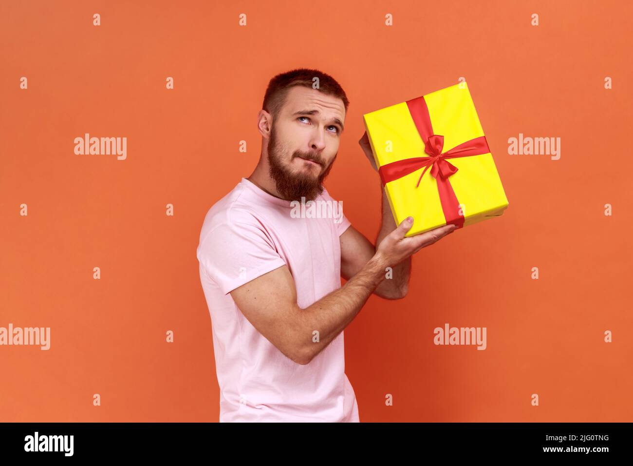 Porträt eines neugierigen bärtigen Mannes, der steht, die gelbe Geschenkbox hält, mit nachdenklichen Ausdrücken wegschaut, denkt, was drinnen, trägt rosa T-Shirt. Innenaufnahme des Studios isoliert auf orangefarbenem Hintergrund. Stockfoto