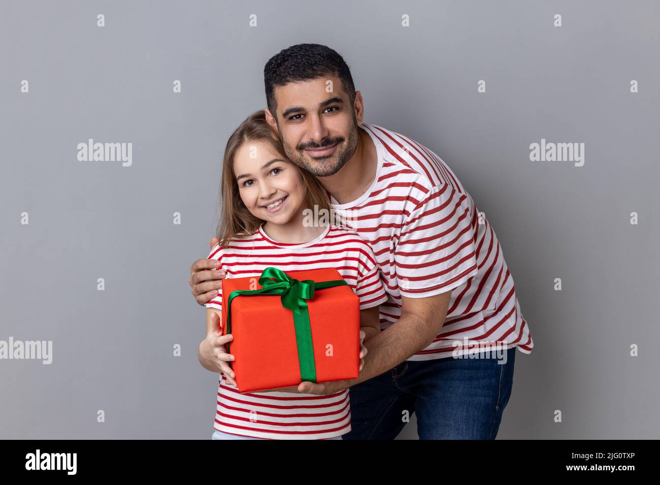 Porträt einer positiven Familie mit rotem Geschenkkarton in den Händen, Vater und Tochter schenken zum Geburtstag der Mutter, zum Ausdruck bringen Glück. Innenaufnahme des Studios isoliert auf grauem Hintergrund. Stockfoto