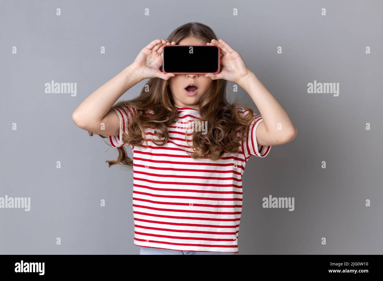 Kleines Mädchen in T-Shirt, die Augen mit Handy bedeckt, posiert mit weit geöffnetem Mund vor Schock Überraschung, unbekanntes Kind versteckt Gesicht mit Handy. Innenaufnahme des Studios isoliert auf grauem Hintergrund. Stockfoto