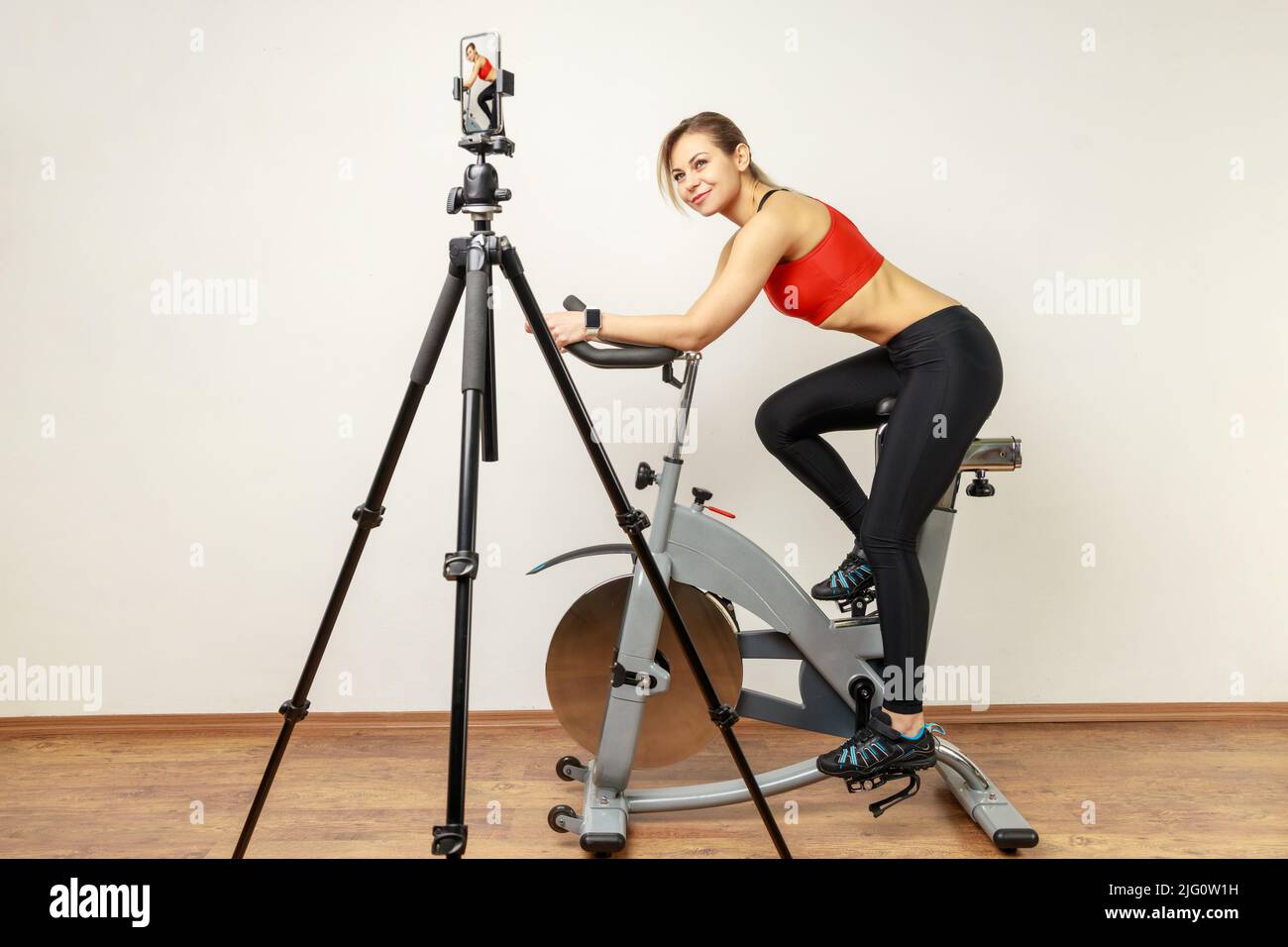 Seitenansicht Porträt einer Bloggerin, die ein Video für ihren Sport-Vlog aufzeichnete, mit Mobiltelefon und Stativ, mit Sportstrumpfhose und rotem Oberteil. Innenstudio auf grauem Wandhintergrund aufgenommen. Stockfoto