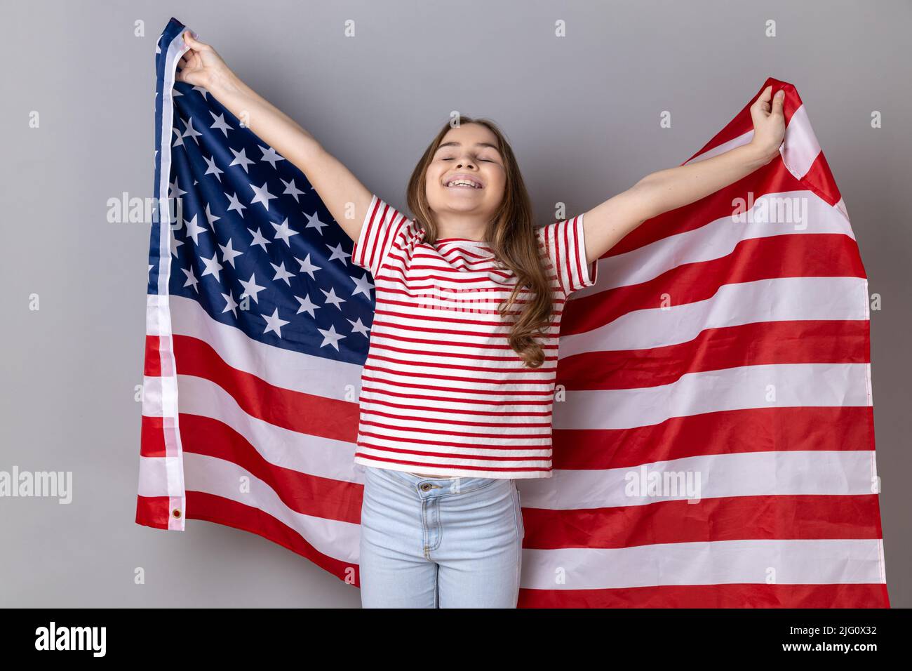 Porträt eines extrem glücklichen kleinen Mädchens mit gestreiftem T-Shirt, das die USA-Flagge über den Schultern hält und die Augen geschlossen und fröhlich lächelt. Innenaufnahme des Studios isoliert auf grauem Hintergrund. Stockfoto