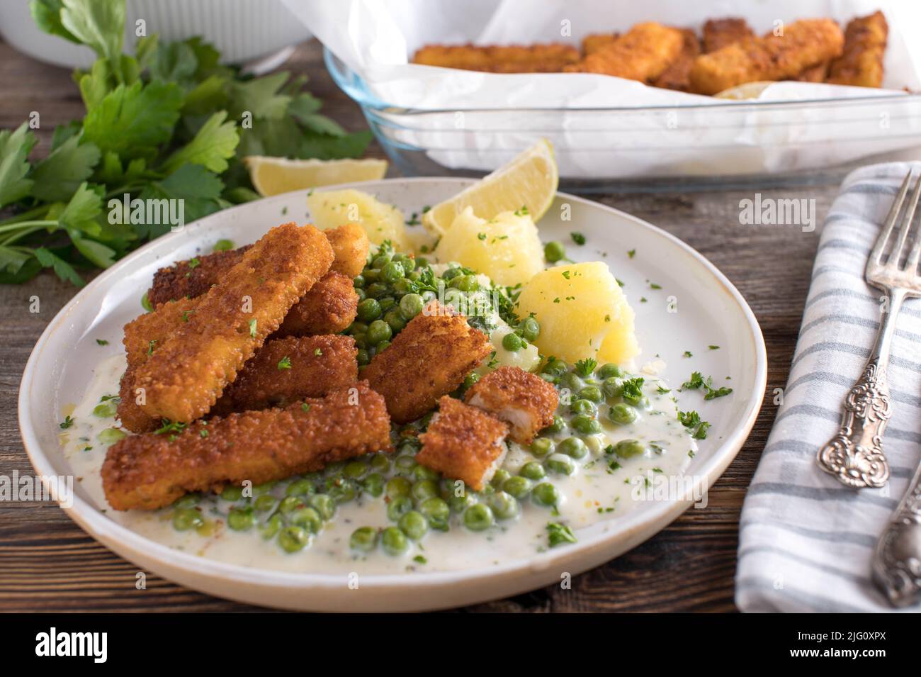 Fischstäbchen mit grünen Erbsen, gekochten Kartoffeln und Bechamelsoße. Serviert auf einem weißen Teller auf einem Holztisch Stockfoto