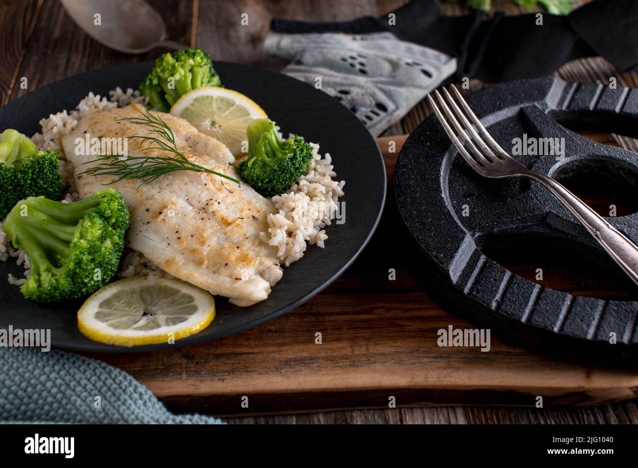 Muskelaufbau Mahlzeit mit gebratenen Fischfilet, braunem Reis und Brokkoli. Serviert auf einem dunklen Teller mit Hantel auf einem Holztisch Stockfoto