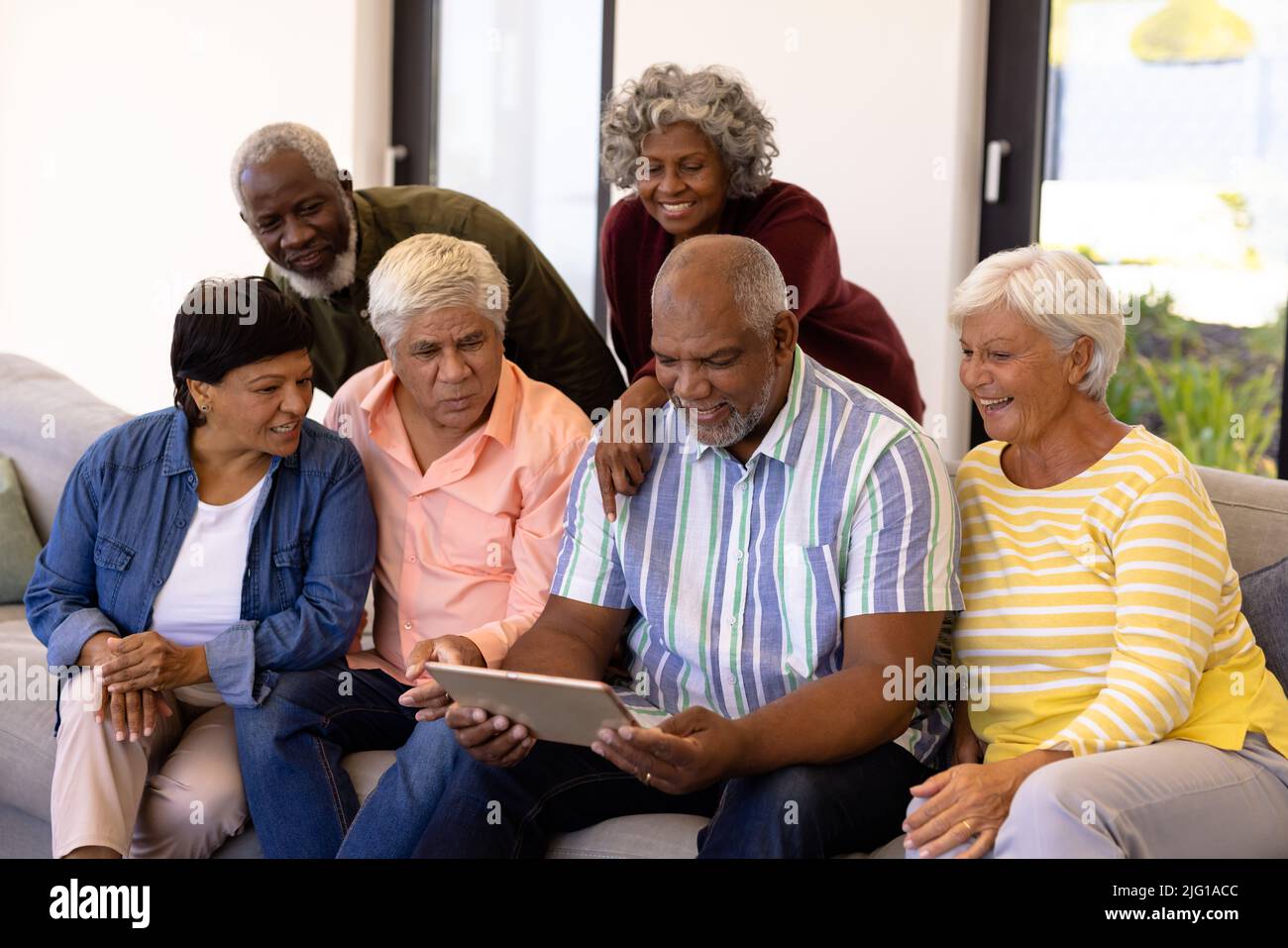Multirassische neugierige Senioren, die einen Freund mit einem digitalen Tablet betrachten, während sie auf dem Sofa sitzen Stockfoto