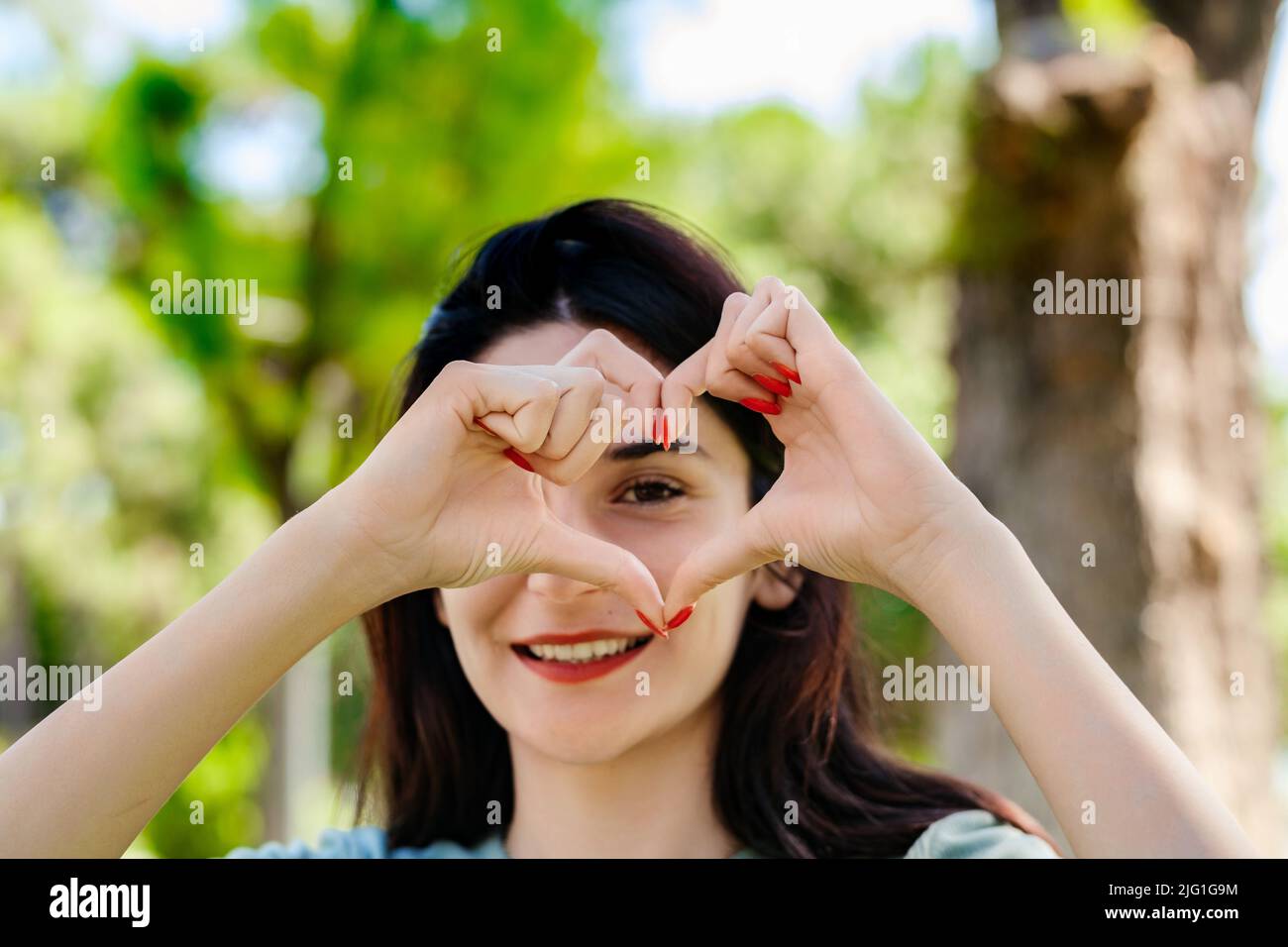 Glückliche Brünette Frau trägt türkisfarbenes T-Shirt im Stadtpark, im Freien tun Herzform mit Hand und Finger lächelnd Blick durch Zeichen Stockfoto
