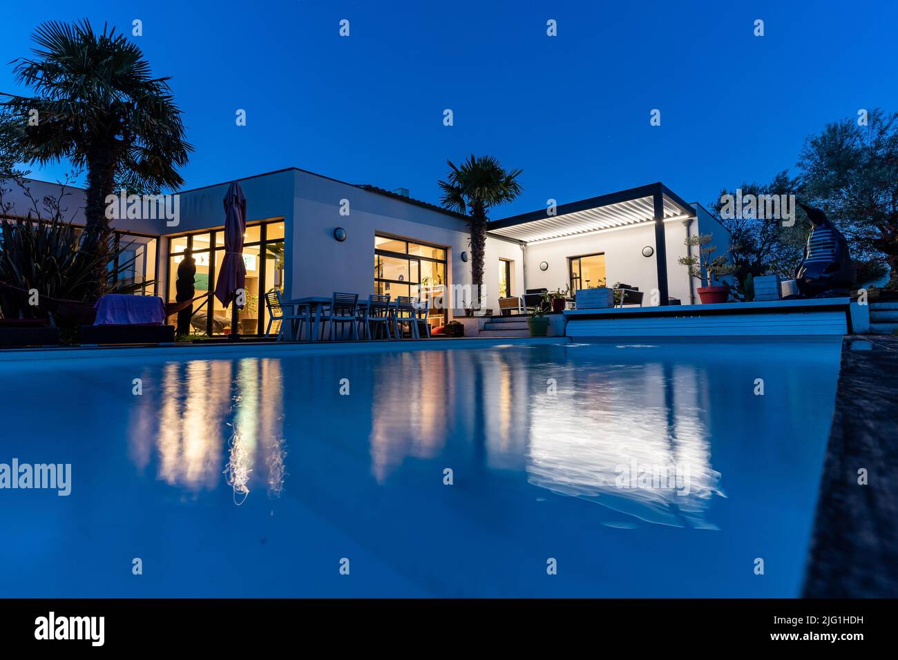 Trendige Terrasse Pergola Schatten Struktur, Markise und Terrasse Dach, Gartenlounge, Stühle, Metall-Grill umgeben von Landschaftsgestaltung. Nachtaufnahme Stockfoto