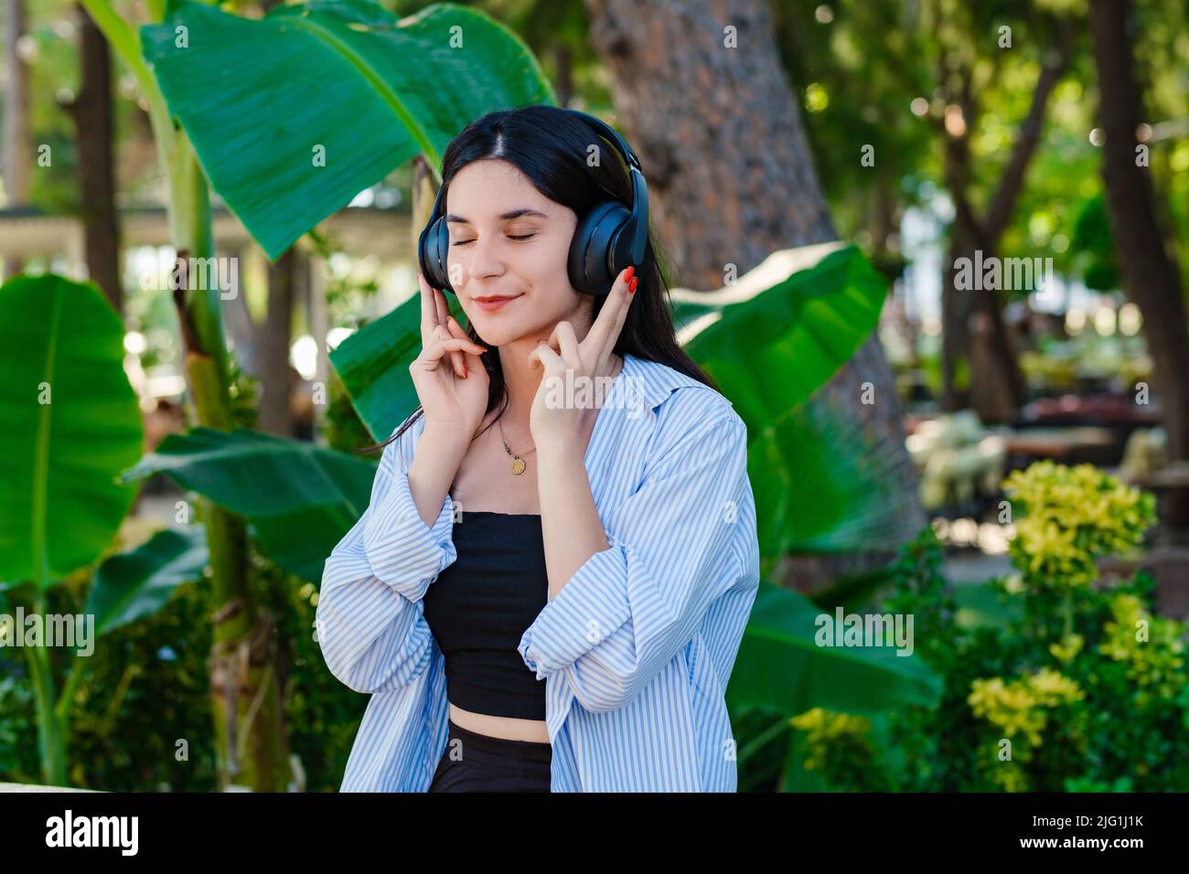 Brünette tausendjährige Frau trägt gestreiftes Hemd stehen auf Stadtpark, im Freien Musik in Kopfhörern und Augen geschlossen hören. Genießen Sie gute Qualität soun Stockfoto