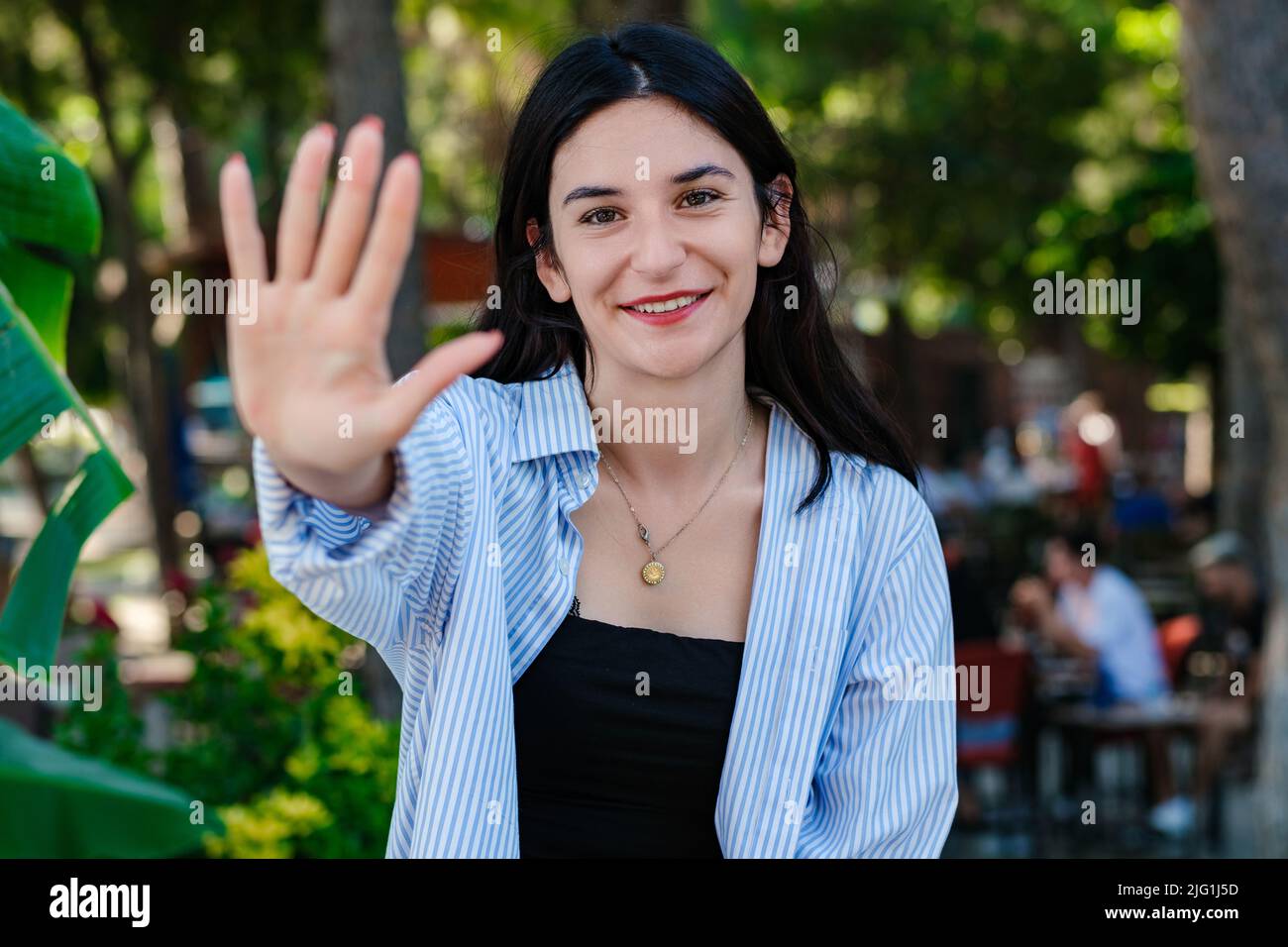 Fröhliche Frau im gestreiften Hemd, die im Stadtpark steht und mit den fünf Fingern im Freien zeigt und nach oben zeigt, während sie selbstbewusst und glücklich lächelt. Stockfoto