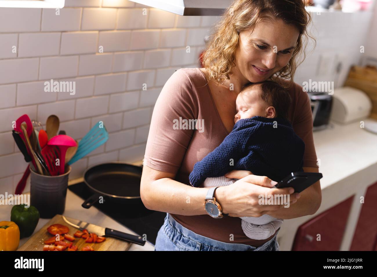 Kaukasische Mutter mit mittlerem Erwachsenen und Smartphone, während sie ein süßes, schlafend neugeborenes Baby in der Küche trägt Stockfoto