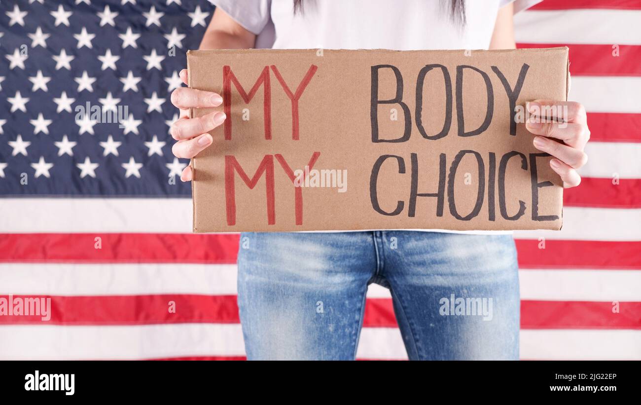Junge Frau Protesterin hält Karton mit My Body My Choice Zeichen gegen USA Flagge auf Hintergrund. Mädchen protestiert gegen Anti-Abtreibungsgesetze. Feministisch Stockfoto