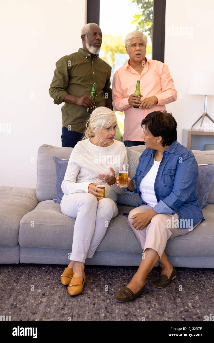 Mehrrassische ältere Frauen mit Bier reden, während Männer im Pflegeheim das Spiel ernsthaft beobachten Stockfoto