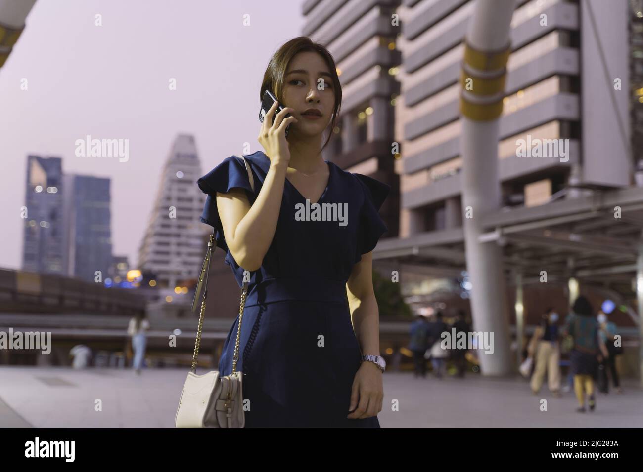 Erfolgreiche und intelligente junge asiatische Frau in legerer Kleidung wartet auf jemanden und telefoniert in der Stadt Straße während des Abends Stockfoto