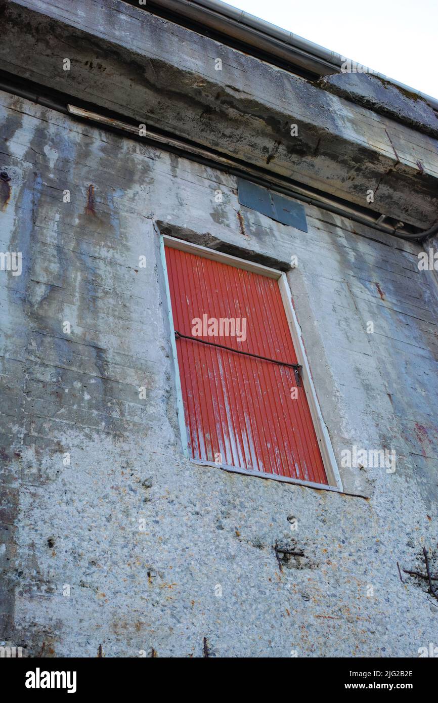 Ein Steingebäude mit einer roten Tür auf einem alten einsamen Grundstück. Architekturdetails des Äußeren einer grauen, industriell gebauten Struktur. Verwittert Stockfoto