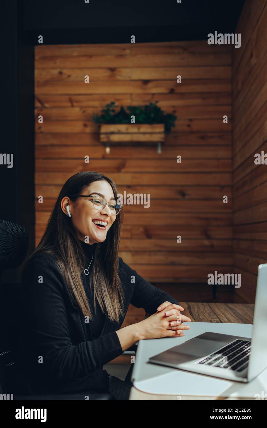 Geschäftsfrau lächelt fröhlich während eines Online-Meetings in einem modernen Co-Working-Raum. Fröhliche Unternehmerin, die mit ihrem Unternehmen kommuniziert Stockfoto
