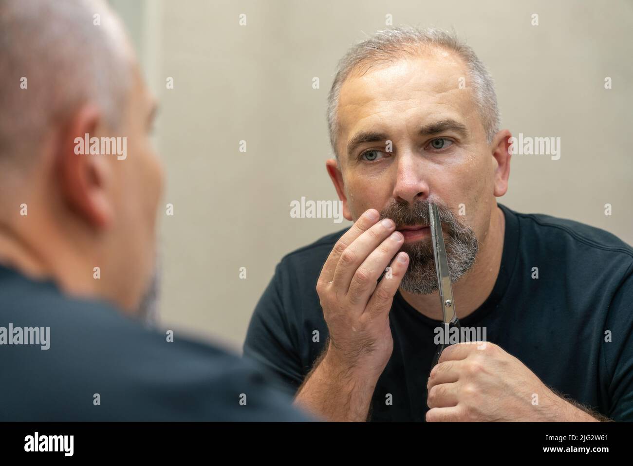 Ein gutaussehender Mann mittleren Alters, der mit einer Schere seinen Bart ein bisschen abschneidet und die Form fixiert Stockfoto