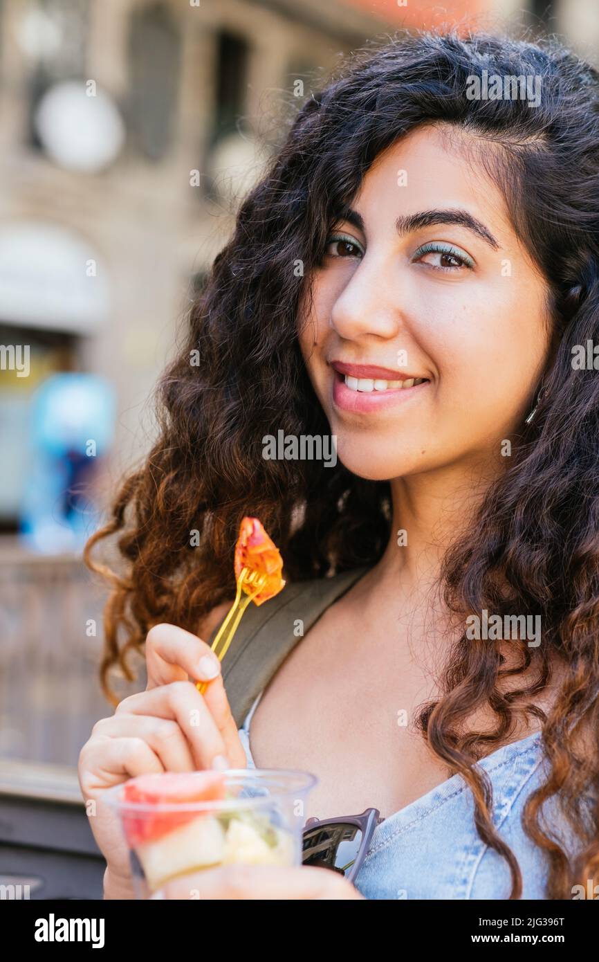 türkische junge Frau, die die Kamera anschaut, während sie einen Plastikbecher mit Fruchtstücken in der Hand hält Stockfoto
