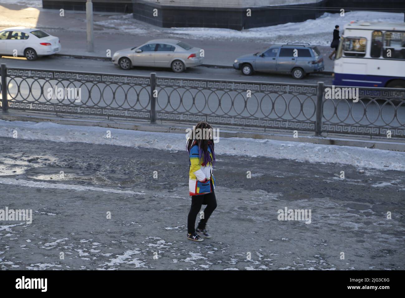 Das Mädchen mit Zöpfen, das einen gelb-blau-weißen Mantel trägt, läuft allein mit Autos, die von einem Zaun getrennt sind, auf einer verschneiten Straße entlang Stockfoto