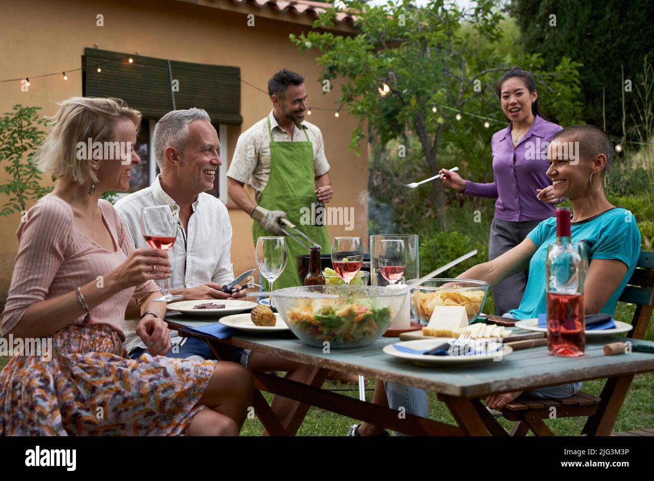 Menschen mittleren Alters essen zu Abend und trinken gemeinsam im Garten Rotwein. Glückliche Freunde essen bbq-Essen im Hinterhof. Ein Lebensstil der Freundschaft Stockfoto