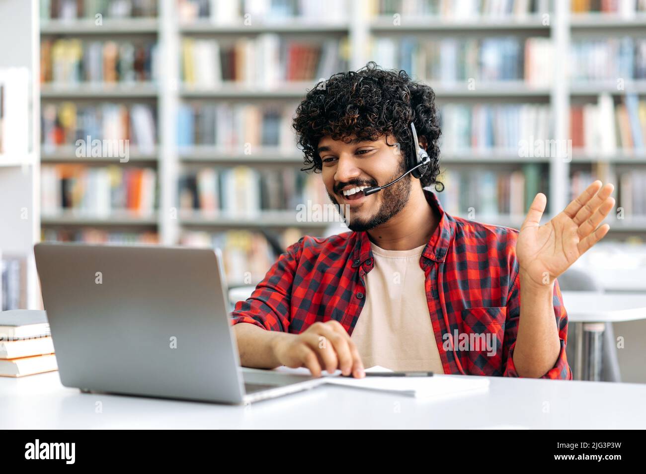 Fröhlicher, positiver indischer oder arabischer Student oder Freiberufler mit Headset, sitzend am Tisch in einer Bibliothek, mit Laptop, im Videoanruf sprechend, winkend, Online-Webinar hörend, Vortrag, lächelnd Stockfoto
