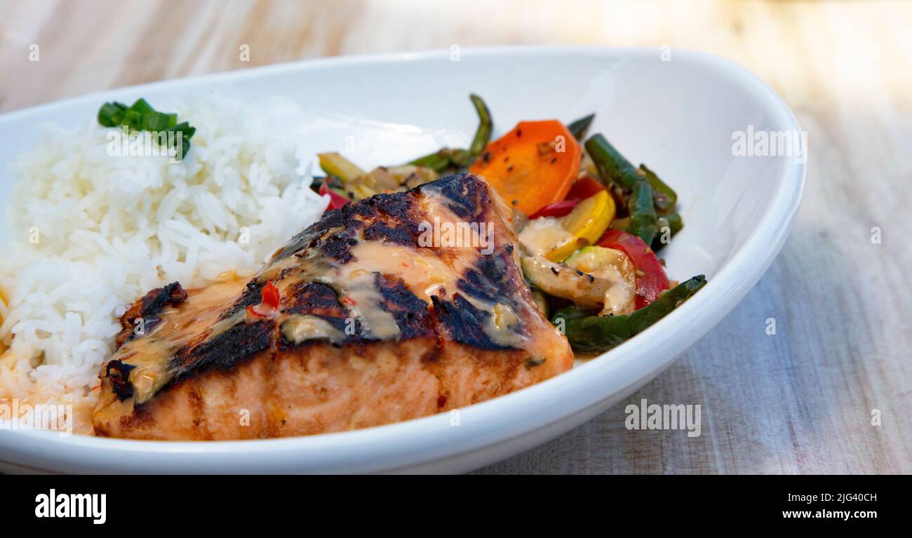 Verkohlter, marinierter Lachs mit Jasminreis und frischem, gesundem Gemüse auf einer weißen Platte auf hellem Holzhintergrund Stockfoto