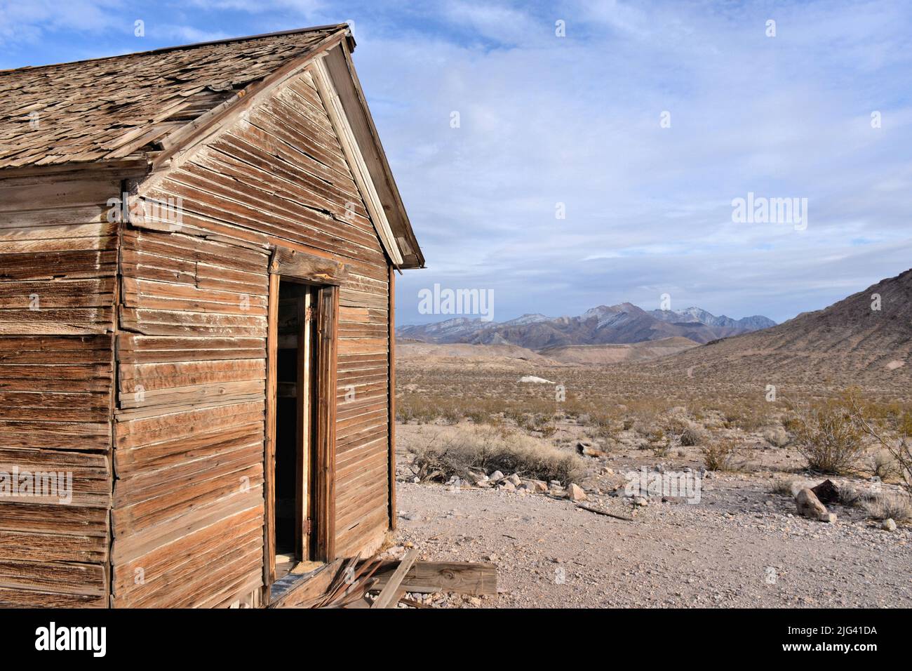 Eine verlassene Holzhütte steht in der Wüste unter einem blauen Himmel, der von Wolken durchsetzt ist. Schnee Flecken Berge in der Ferne. Stockfoto