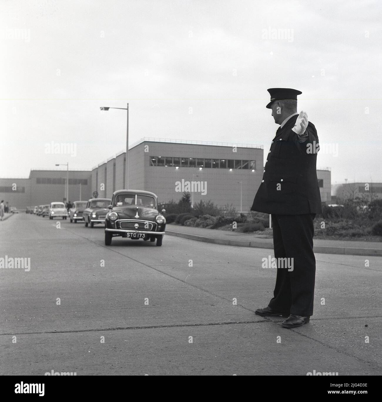 1950s, ein historischer, uniformierter Sicherheitsmann, der die Autos signalisiert, die das riesige Stahlwerk in Port Talbot, Wales, Großbritannien verlassen. Stockfoto