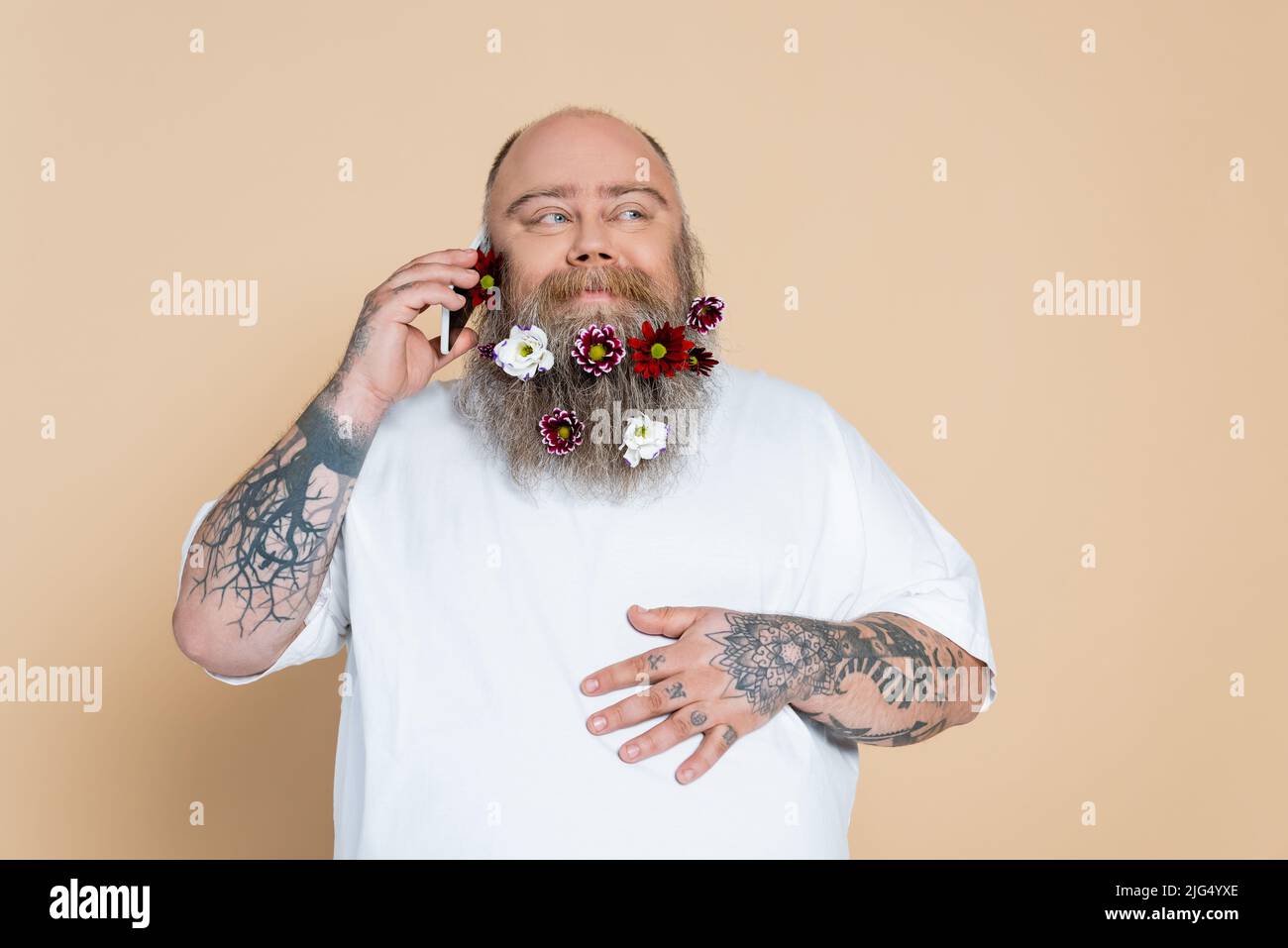 Fröhlicher Mann mit Blumen im Bart, der auf einem Mobiltelefon auf Beige isoliert spricht Stockfoto