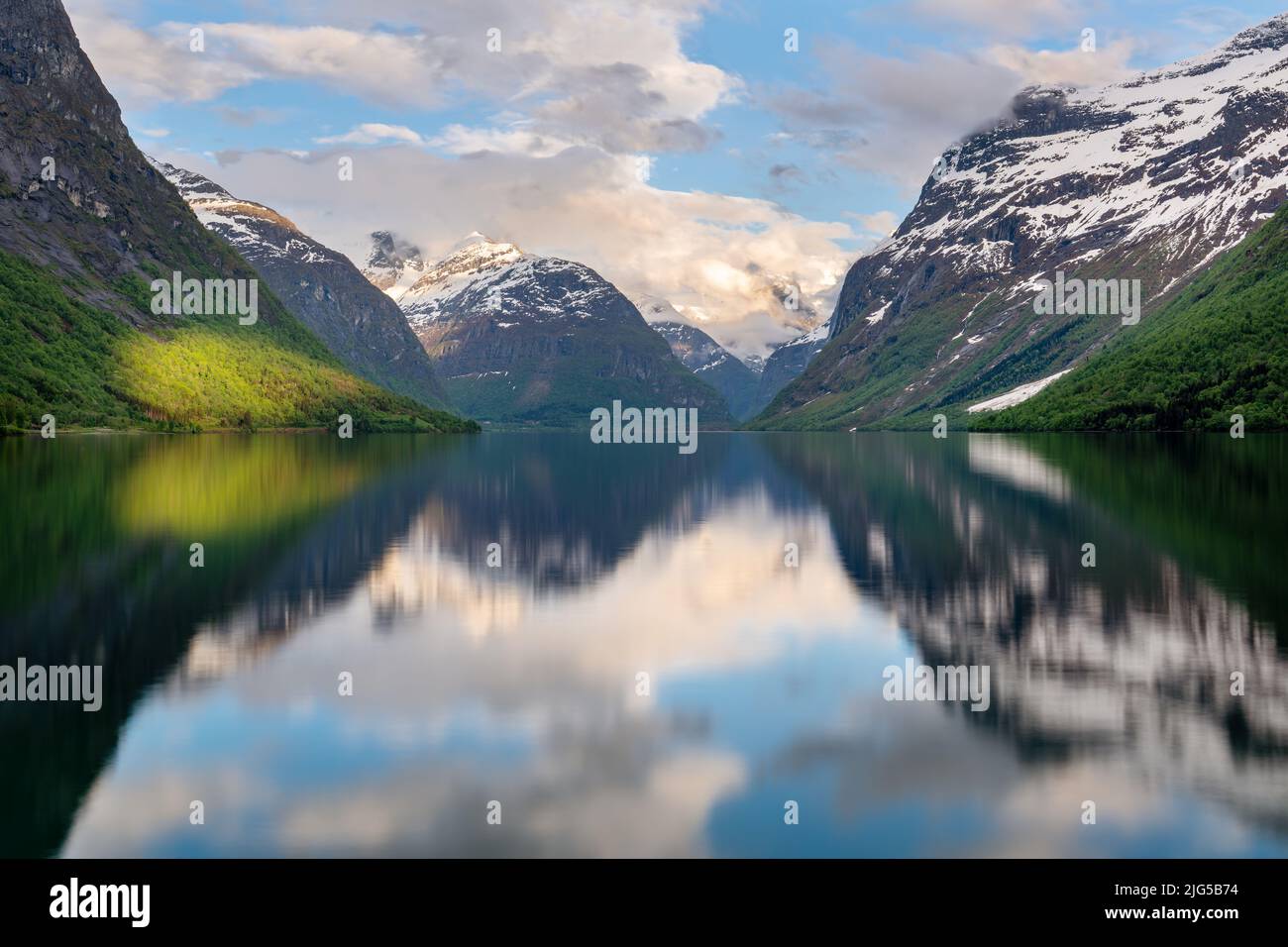 Blick über einen See umgeben von hohen, teilweise schneebedeckten Bergen in Norwegen, die Sonne erhellt auffallend ein Gebiet am Ufer, Spiegelung Stockfoto