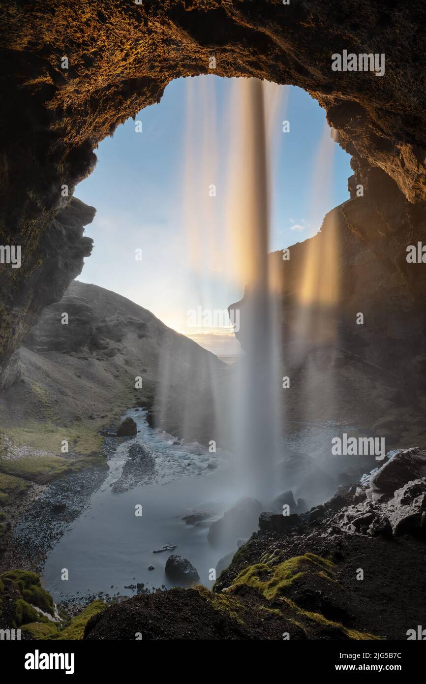 Blick durch einen Wasserfall bei Sonnenuntergang in Island, die untergehende Sonne färbt das Wasser, der Blick deckt auch den Bereich vor dem Wasserfall ab Stockfoto