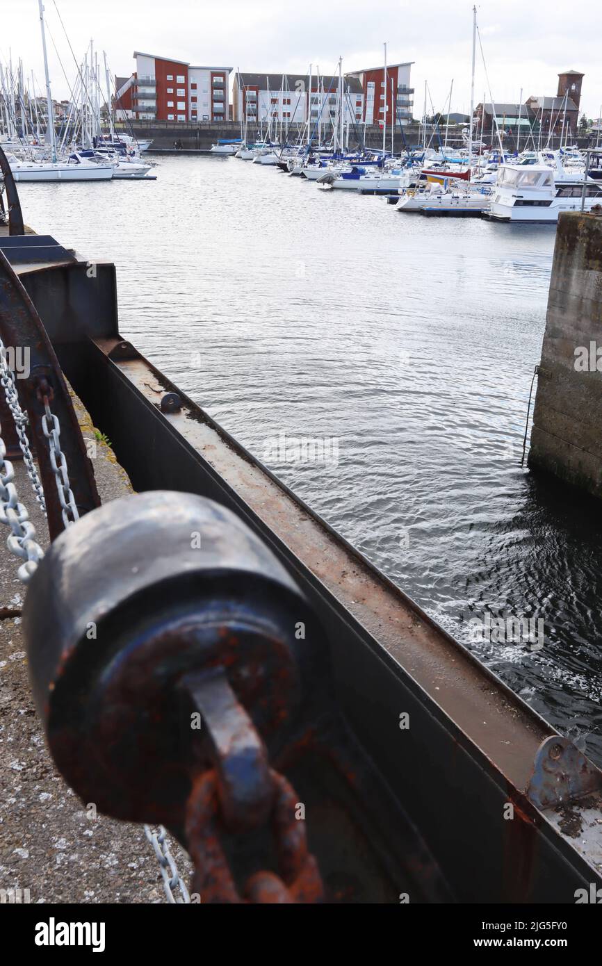 Eine rostige Kette sichert einen Betonpfoller mit Booten, die im Hintergrund an einem modernen Yachthafen angedockt sind Stockfoto