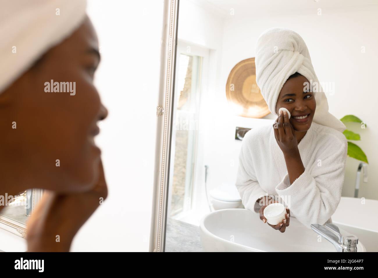 Lächelnde junge afroamerikanische Frau, die ein Beauty-Produkt anwendet, während sie im Spiegel auf das Badezimmer schaut Stockfoto
