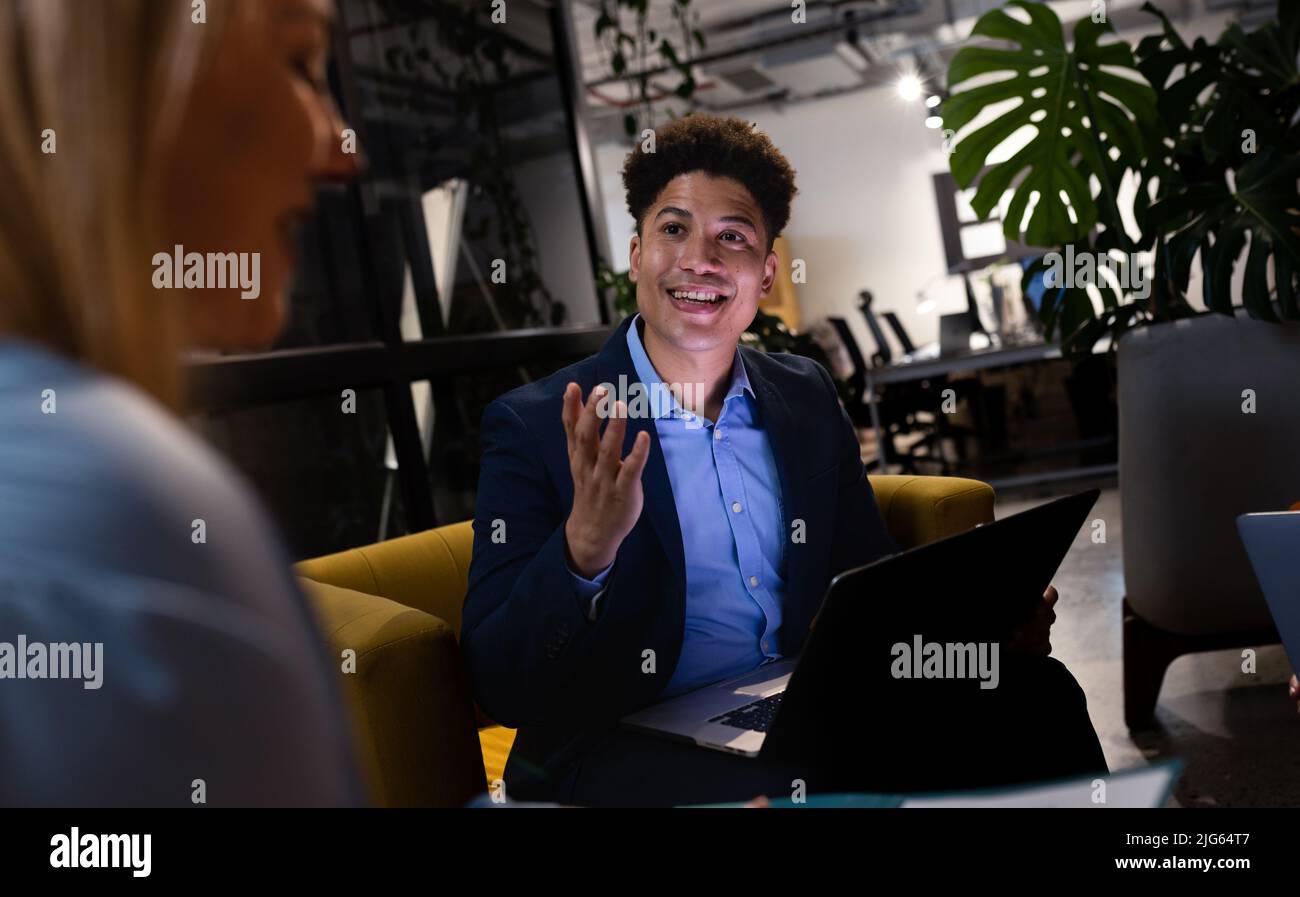 Lächelnder asiatischer Geschäftsmann, der über einen Laptop arbeitet und mit einer kaukasischen Kollegin im Büro spricht Stockfoto