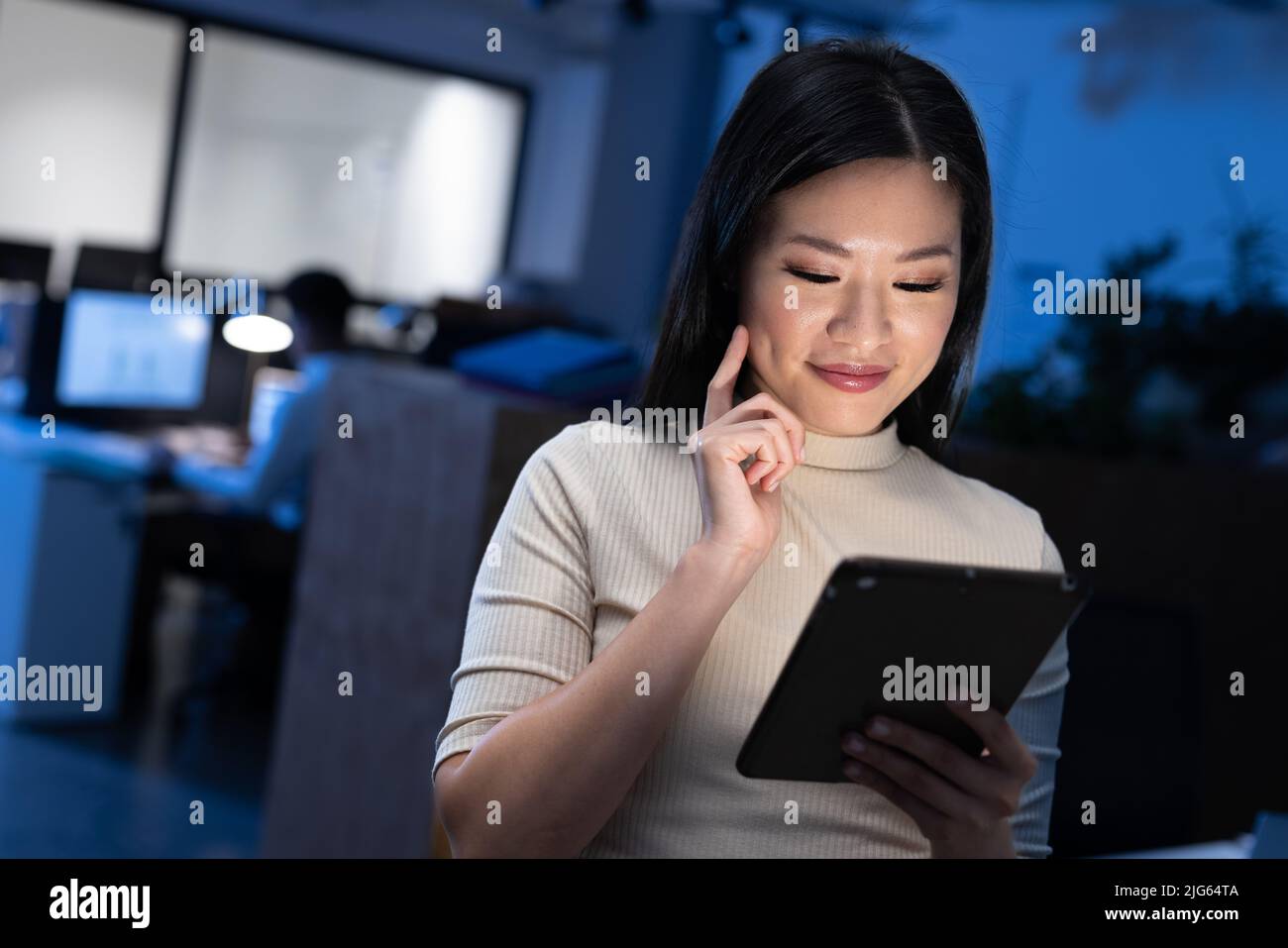 Lächelnde asiatische Geschäftsfrau mit digitalem Tablet, während sie spät im Büro in der Nacht arbeitet Stockfoto