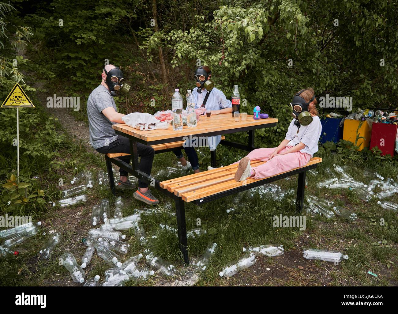 Mann, Frau und Kind in Gasmasken sitzen am Tisch im Wald mit Müllplastiktüten und giftigen Zeichen. Familie, die im Wald ruht, mit dem Warnsymbol für Abfall und Schädel und Kreuzknochen. Stockfoto