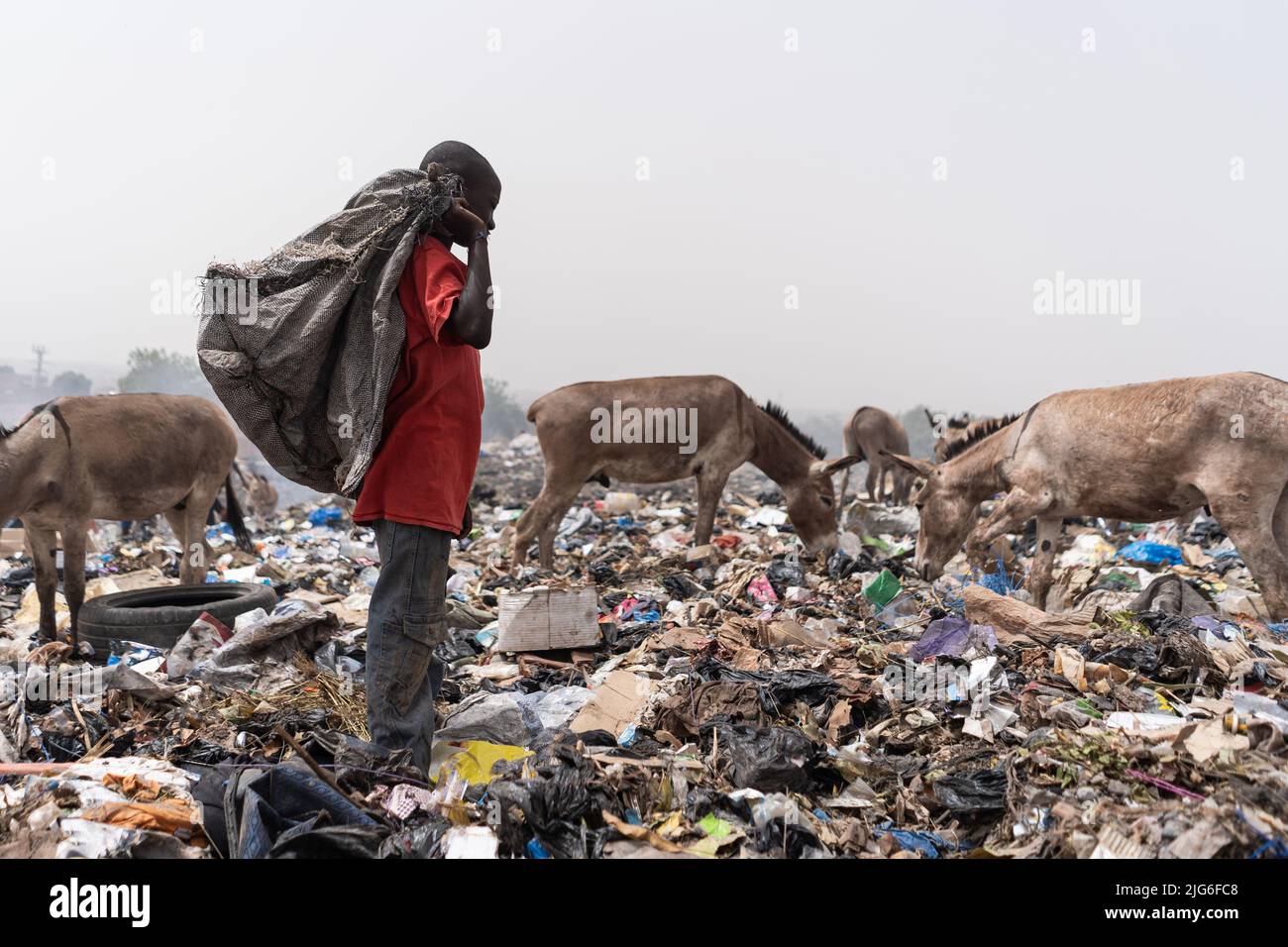 Einsamer arrikanischer Junge, der auf einer Deponie mit einer schwarzen Plastiktüte auf der Schulter auf der Suche nach wiederverwendbarem Material steht, umgeben von hungriger Müllweide Stockfoto