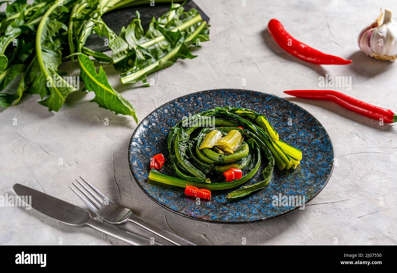 Zichorien-Grüns mit Knoblauch und heißem rotem Papier auf blauem Teller mit Zutaten und Besteck zur Seite geschmort. Stockfoto