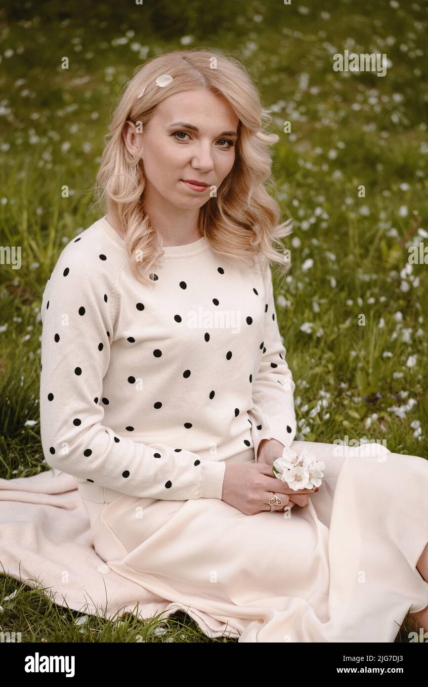 Glückliche blonde Frau, die im weißen Kleid auf dem grünen Gras sitzt und den blühenden Frühling genießt Stockfoto