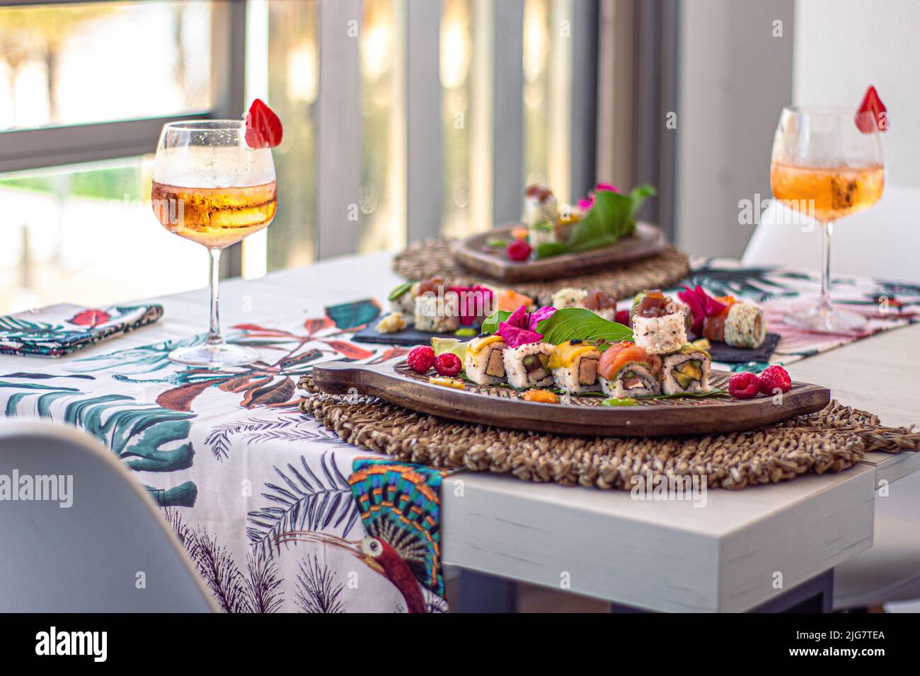 Eine Nahaufnahme von Sushi-Sets auf Tellern auf einem weißen Tisch mit einer tropischen Tischdecke und Tassen Champagner mit verschwommenem Hintergrund Stockfoto