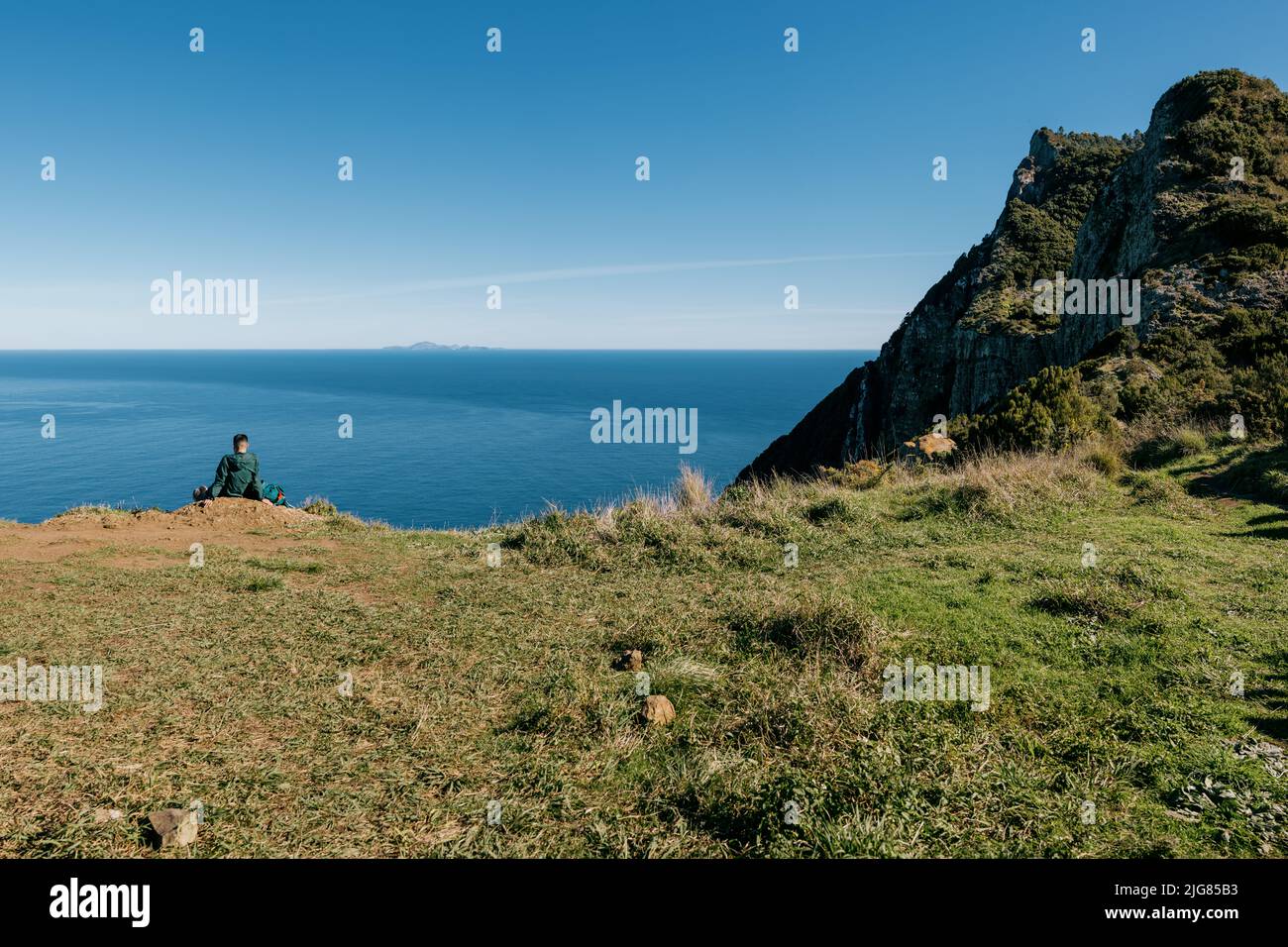 Der Mann, der am Rand der grünen Klippe gegen das blaue Meer und den blauen Himmel sitzt. Madeira, Portugal. Stockfoto